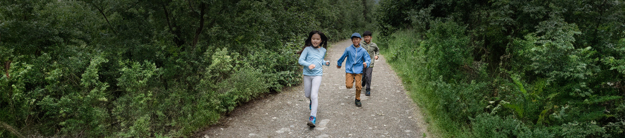 Three children running down a path outside, with many trees in the background.