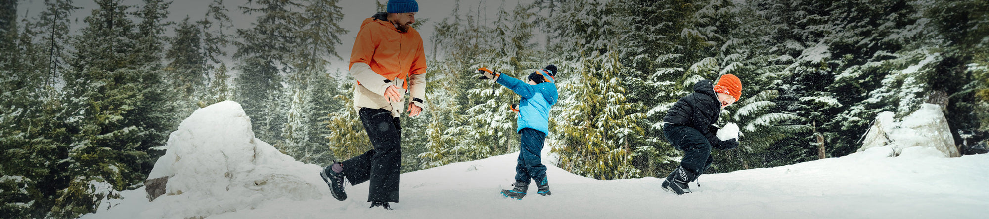 A man and two children playing in the snow, wearing outdoor winter attire, with snow-covered evergreen trees in the background