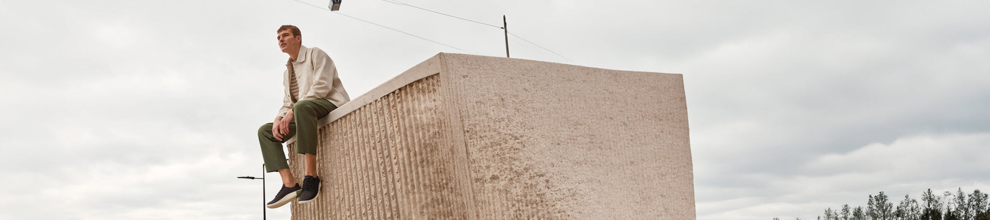 A man sitting at the edge of the top of a building, wearing olive green pants, and cream jacket, and black KEEN slip on shoes