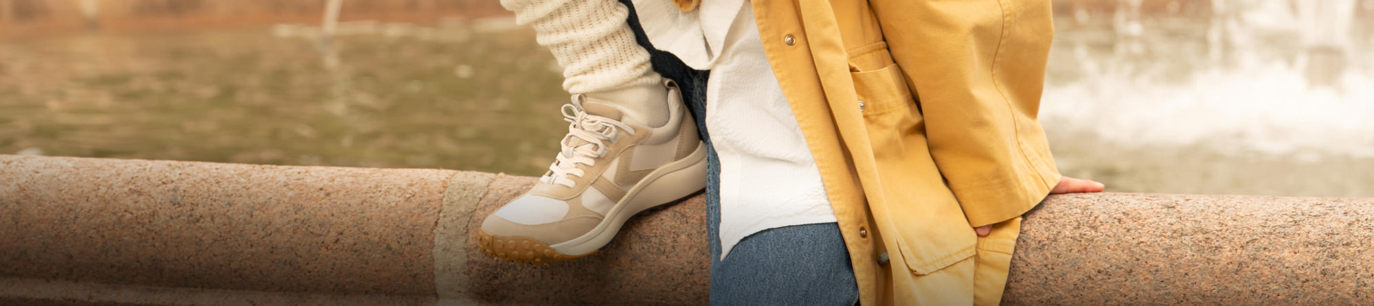 A closeup of someone's shoe up on a ledge. The shoe is a white and beige, casual sneaker, and the person is wearing jeans, a white shirt, and a yellow jacket.