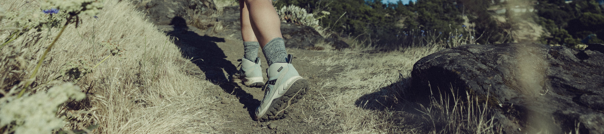 Someone shown from the knees down walking across forest terrain, wearing KEEN hiking sneakers.
