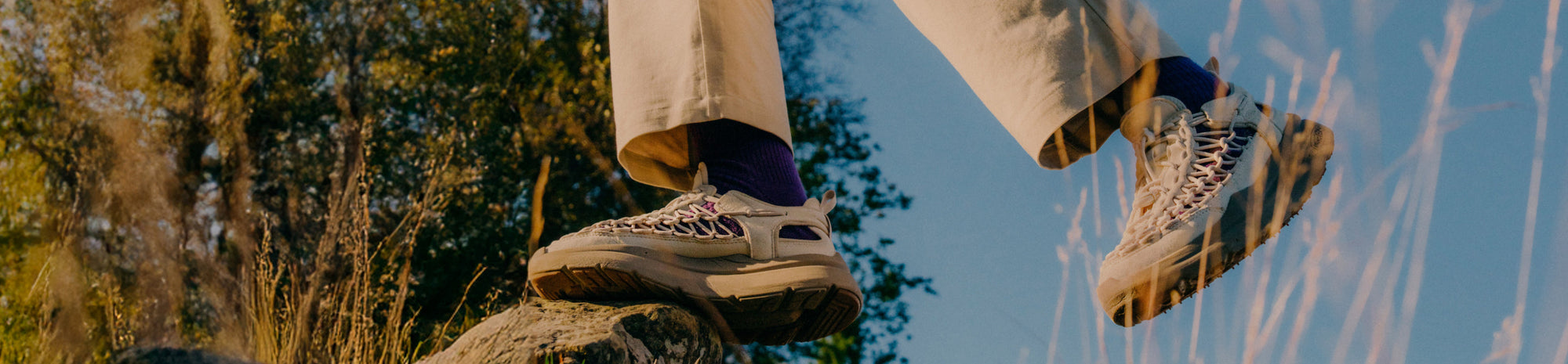 A woman in a black T-shirt and leggings, wearing cream-colored KEEN walking shoes, walking next to what appears to be a strip of store entrances.