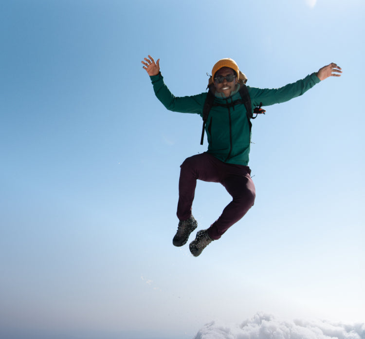 A person wearing outdoor clothing, hiking boots, and a backpack, jumping into the air.
