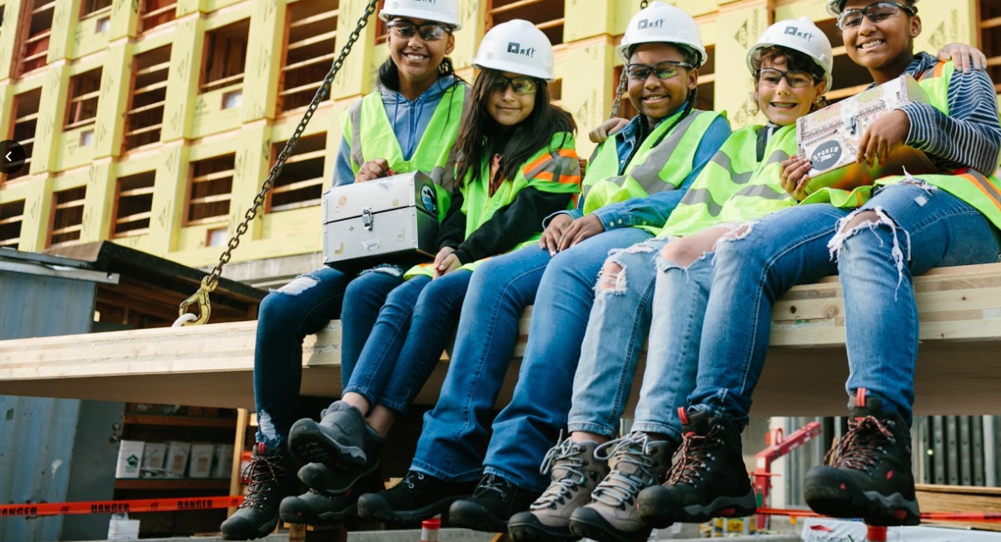 group of five kids wearing jeans, various keen boots, contstructions hats and high visisbility vests sitting on construction site bench
