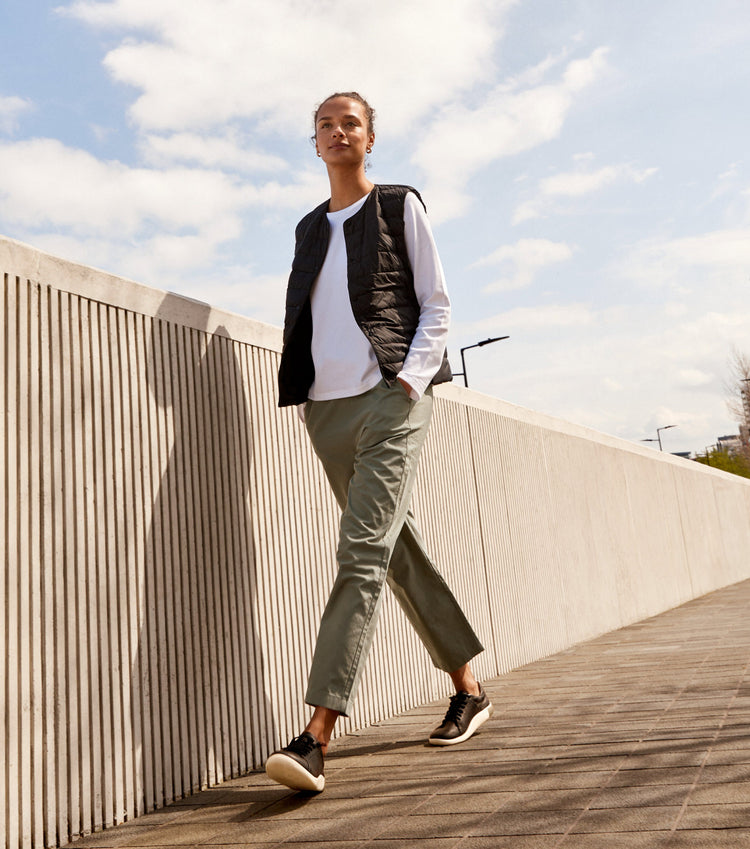 Woman wearing black KNX sneakers and green cargo pants, walking aside a textured cement wall.