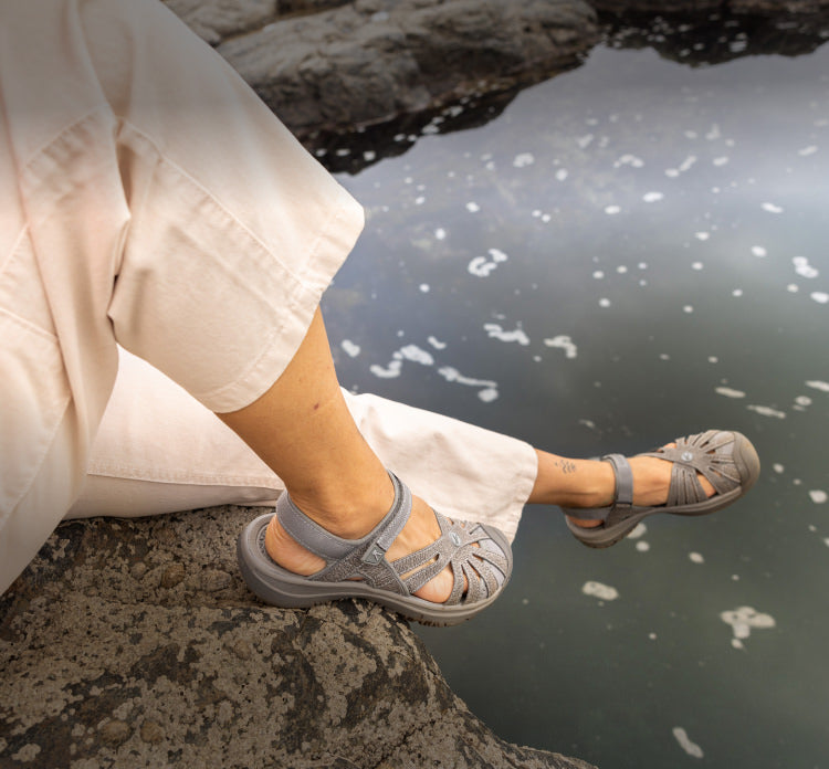 A person sitting on a rock, dangling their legs over a pond, wearing a pair of Rose sandals.