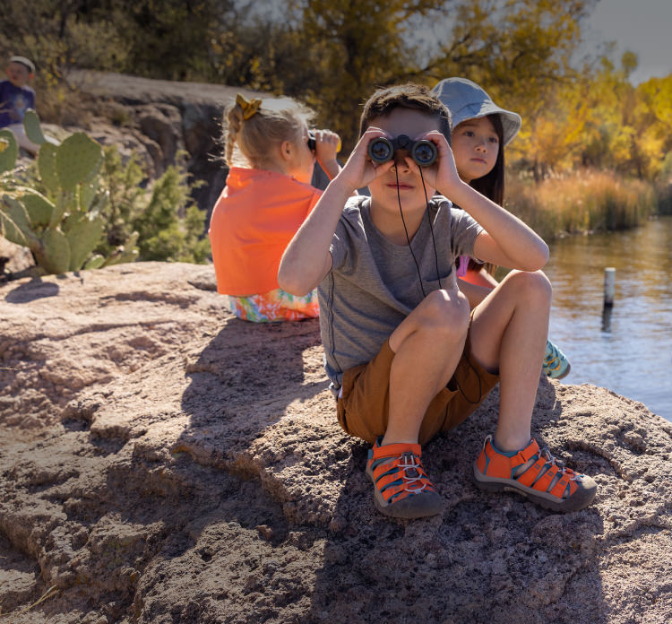 Three children sitting outside by a pond, one wearing binoculars.