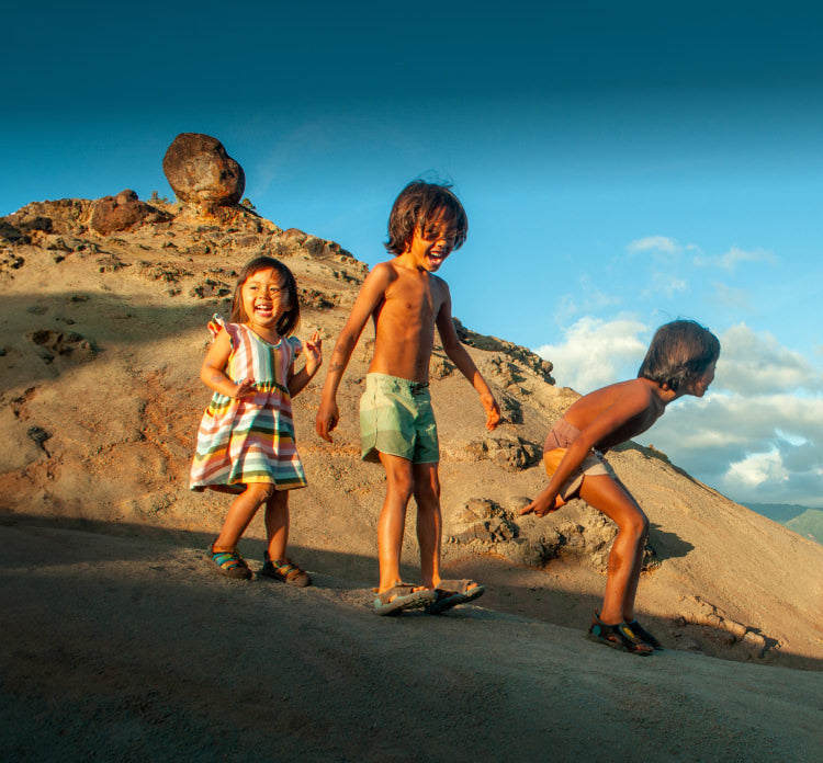 Three young children playing outside in the sand, wearing KEEN kids' sandals.