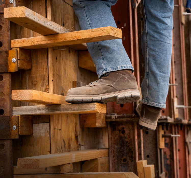 A person wearing brown San Jose work boots and jeans, climbing wooden planks on a job site.