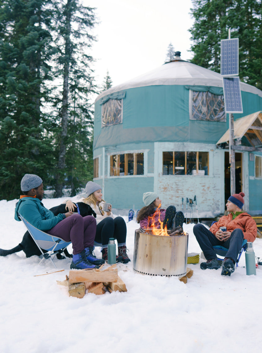 Four people sitting outside in the snow in winter attire, around a fire.
