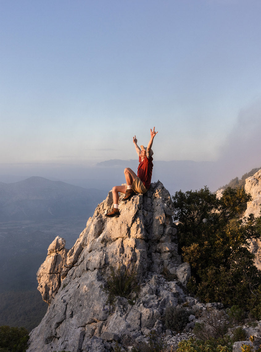 A person sitting at the edge of a cliff, on top of a rock, with their hands up in the setting sun.