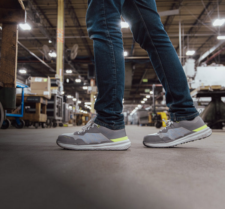 A person wearing grey KEEN work sneakers and jeans in a warehouse.