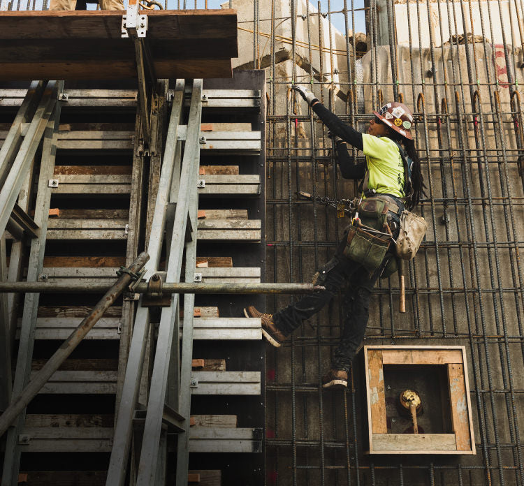 A construction worker climbing scaffolding on a job site, wearing a tool belt and protective gear.
