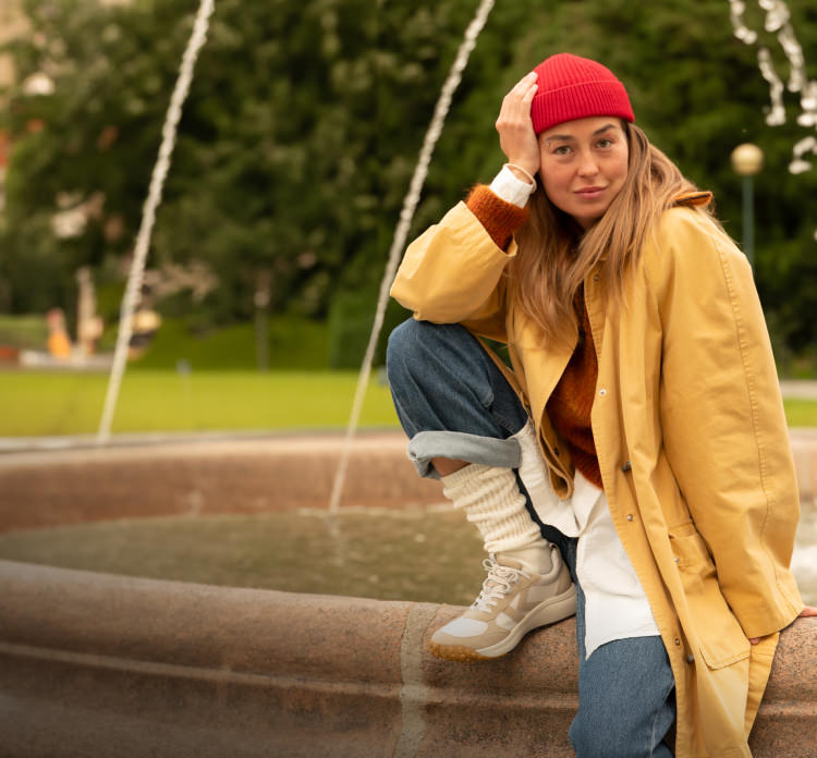 A woman sitting on the edge of a fountain, wearing a jacket, a red beanie hat, and KEEN sneakers.