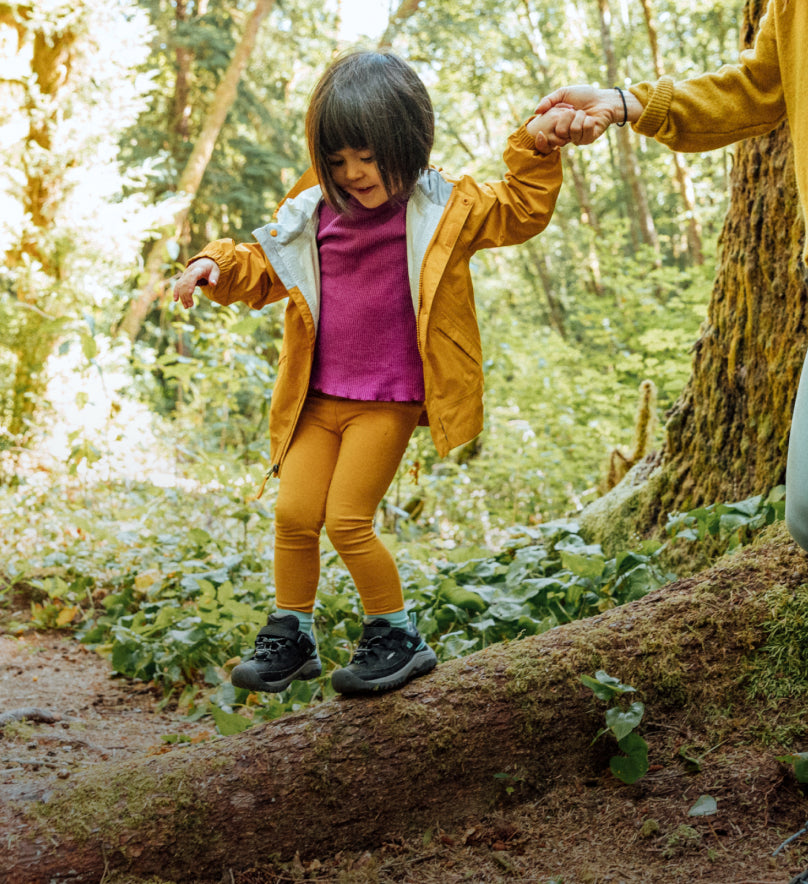 Little girl wearing yellow pants and jacket, holding the hand of an adult while walking across a log wearing her gray Targhee shoes