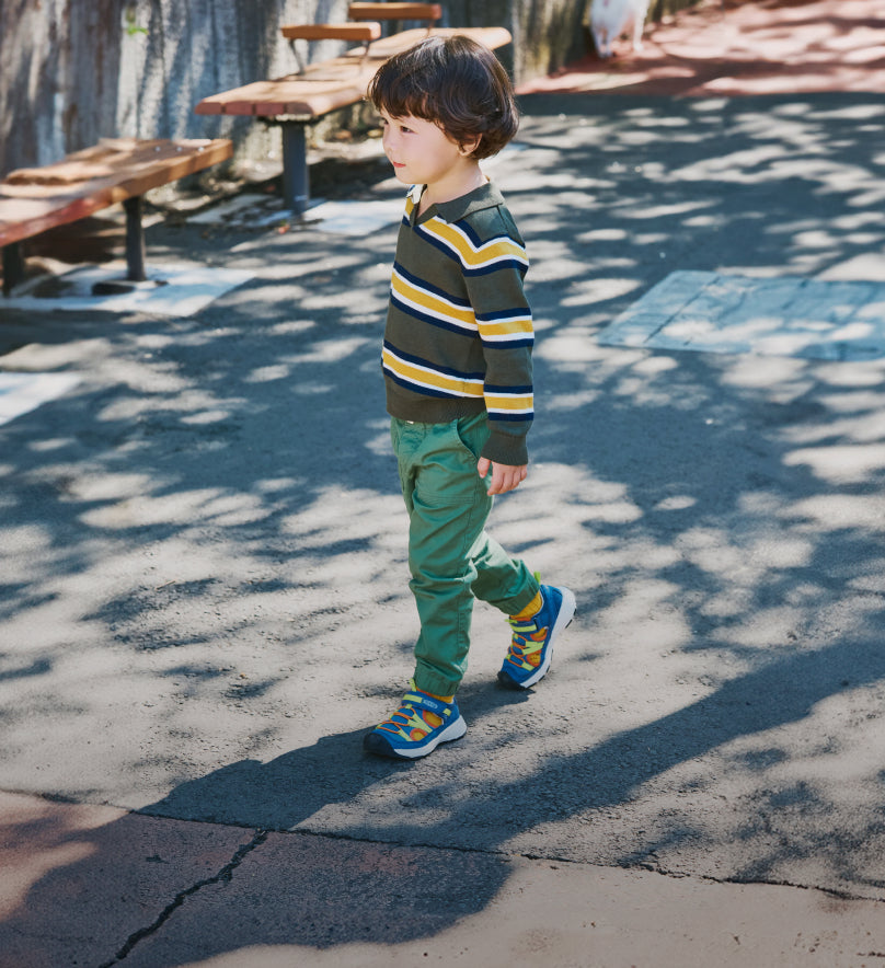 Young boy wearing colorful striped shirt, green pants and blue Motozoa sandals walking along sidewalk. 