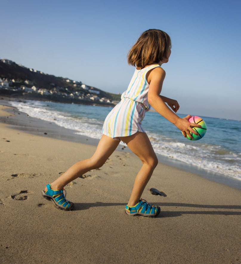 Young girl playing with a bouncy ball on the beach and running in her Newport H2 sandals. 