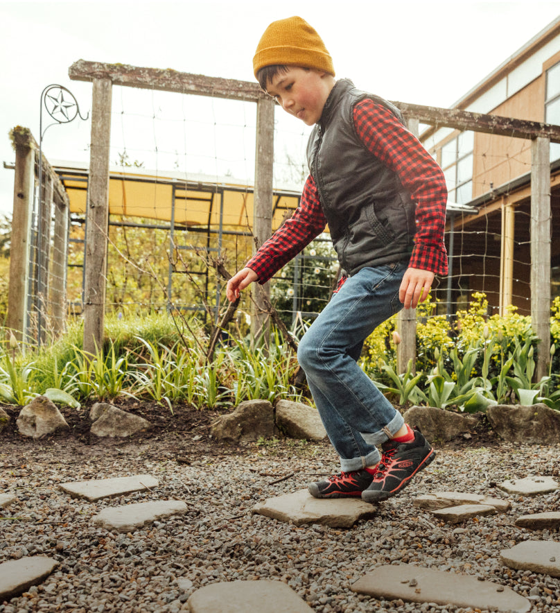Full body shot of boy wearing red and black Chandler 2 sneakers and jumping across stone pavers in a garden. 