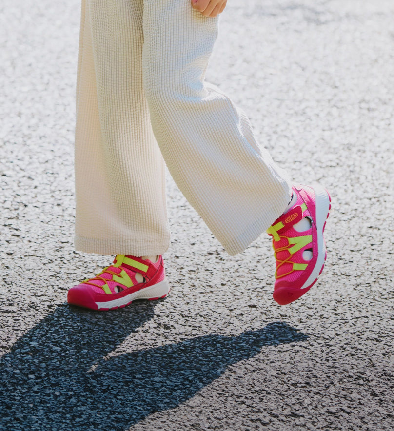 Kneed down shot of child wearing white pants and bright pink and yellow Motoxoa sneakers while walking across pavement. 