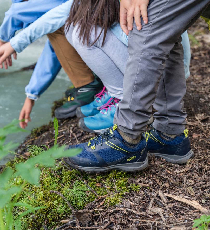 Waist-down shot of three kids wearing Wanduro waterproof hiking shoes and crouching down to point at pond water. 