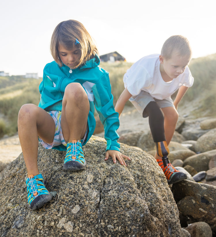 A young girl wearing a blue jacket and a young boy with a bionic leg both wearing Newport Boundless sandals and climbing on beach rocks. 