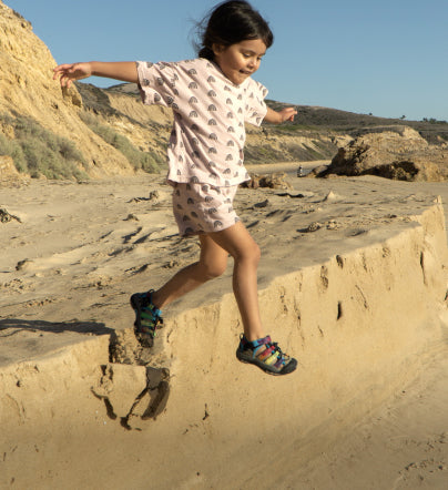 Young girl wearing tie-dye newport h2 sandals and jumping of a sandy ledge at the beach.