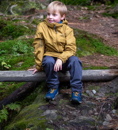Little boy wearing yellow raincoat sitting on a fallen tree and wearing blue and yellow Skua waterproof boots