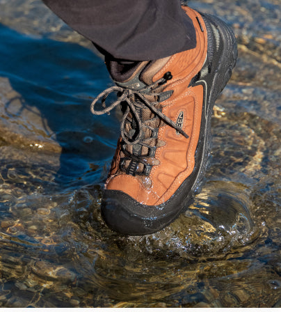 A person wearing a pair of light brown KEEN hiking boots, stepping through water.