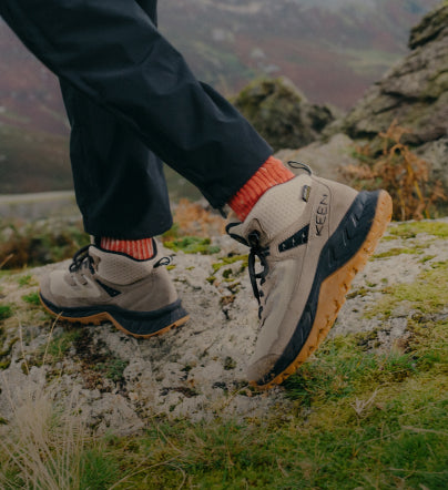 A person wearing a pair of beige KEEN hiking boots, walking across grassy terrain.