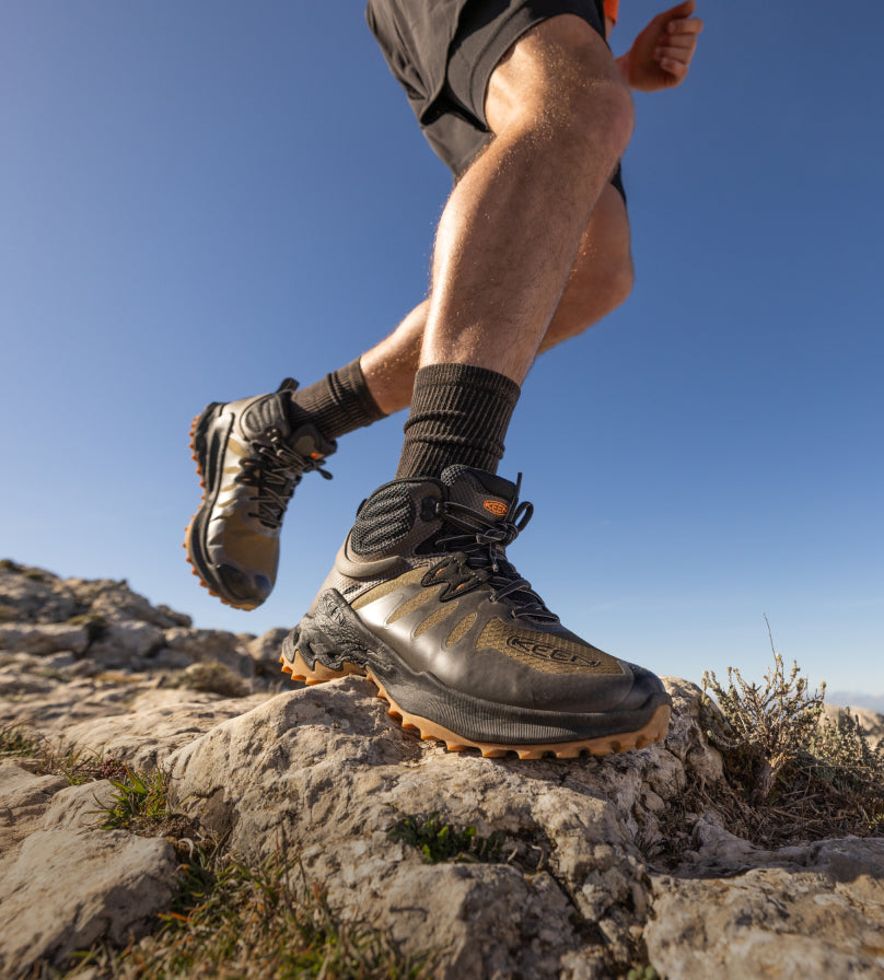 Knee-down shot of man wearing dark green Zionic mid Hiking boots and running across rocky terrain. 