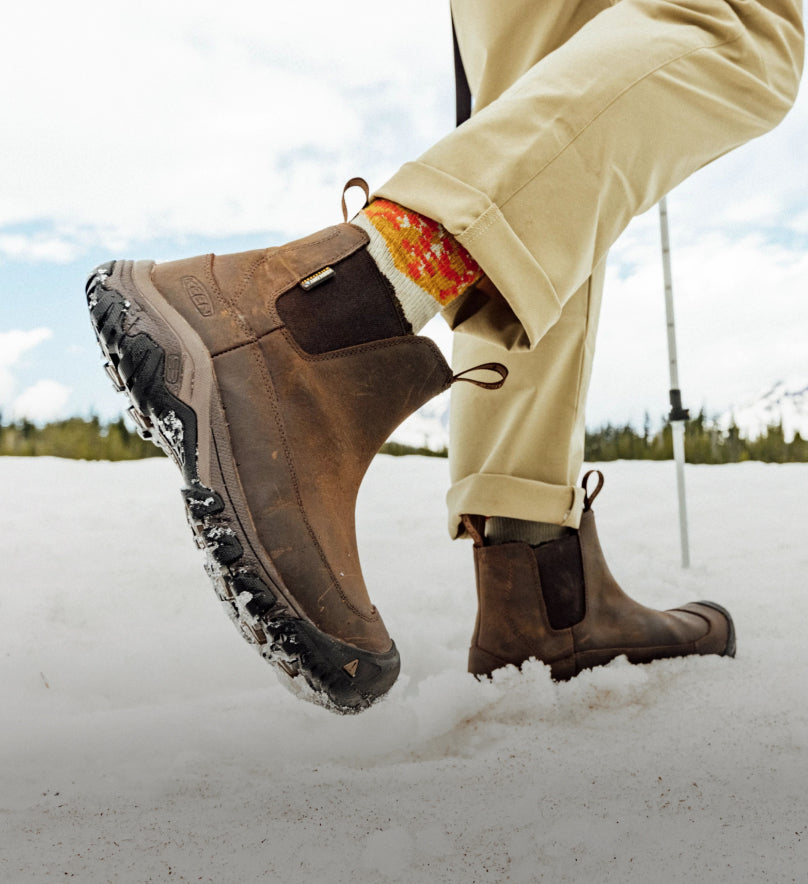 Knee downs shot of man wearing tan pants and brown Anchorage boots while walking across a snowy field. 