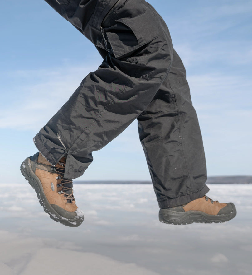 Waist-down shot of man in black snow-pants jumping over icy surface while wearing brown and black Revel IV's.