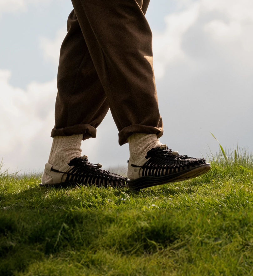 Knee-down shot of man wearing brown pants and brown Uneek sandals with tan socks while walking across a grassy field. 