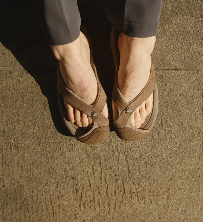 Knee-down shot of man wearing tan Waimea flip-flops against brown, textured cement. 