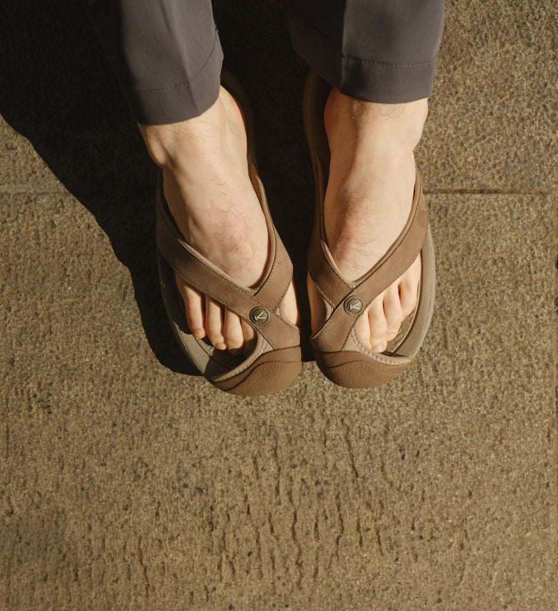 Knee-down shot of man wearing tan Waimea flip-flops against brown, textured cement.