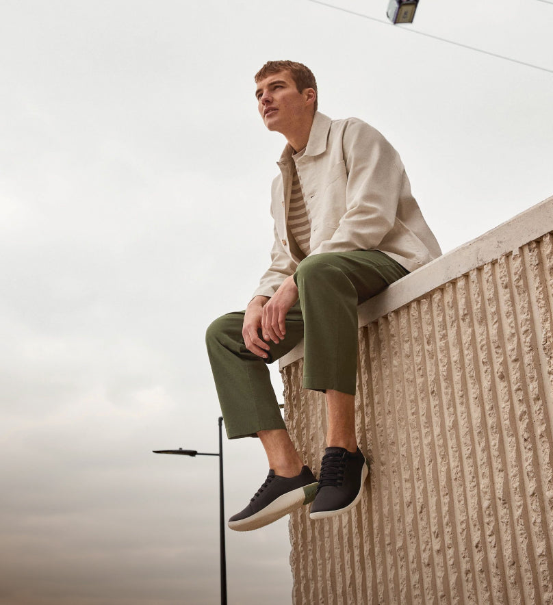 Man wearing black KNX sneakers and olive pants while sitting on textured cement corner ledge. 