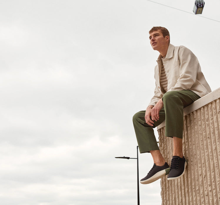 A man sitting on top of a wall, wearing casual sneakers by KEEN.