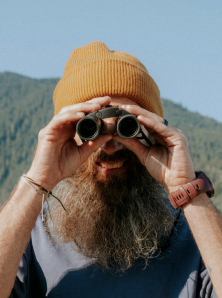 Man with beard and yellow beanie looking through binoculars directly into camera