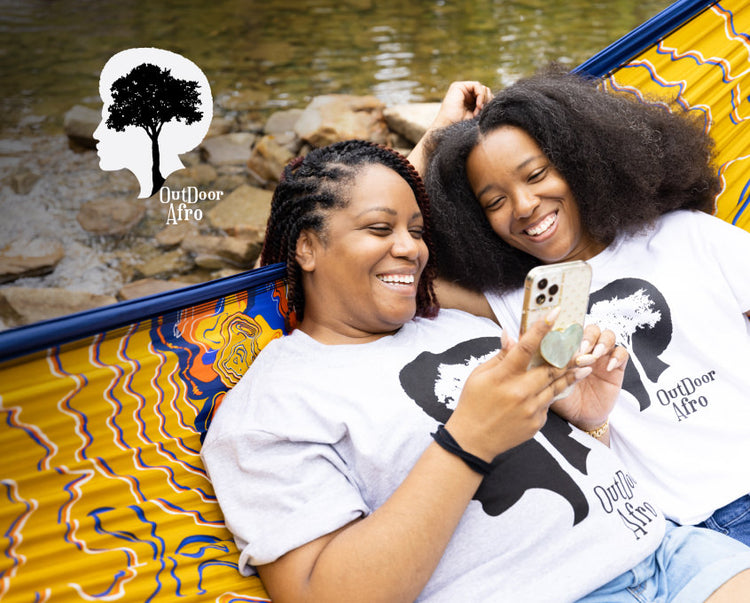 Younger and older woman wearing outdoor afro shirts and sitting together in a hammock, both smiling and watching something on their phone. 