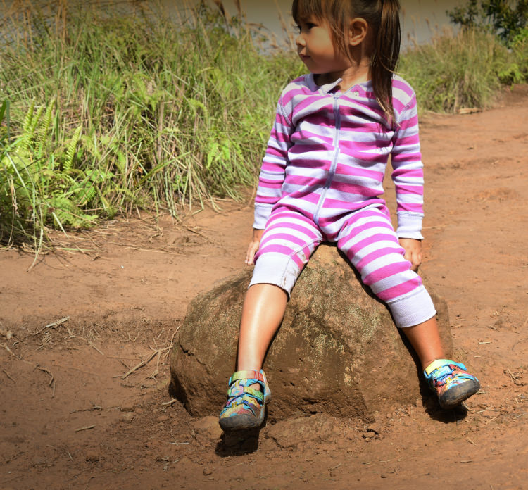 A young girl in a striped-pink onesie, wearing tie-dye KEEN sandals, sitting on a rock outside.