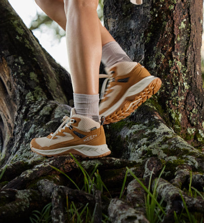A person wearing a pair of orange and white KEEN hiking sneakers, walking over some tree roots.