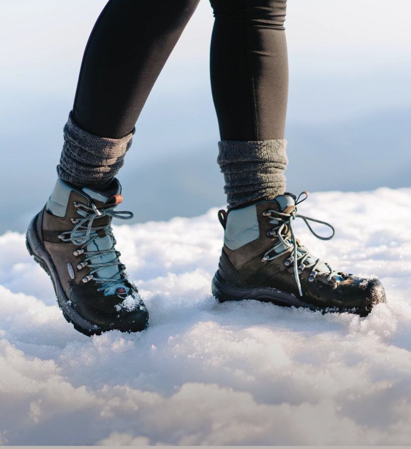 A person wearing a pair of black and blue KEEN snow boots, walking through snow.