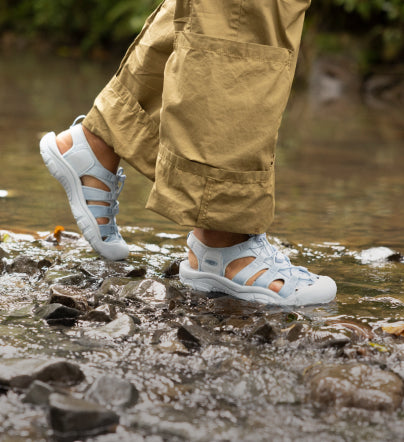 A person wearing light blue Newport H2 sandals on rocky terrain, next to a creek.
