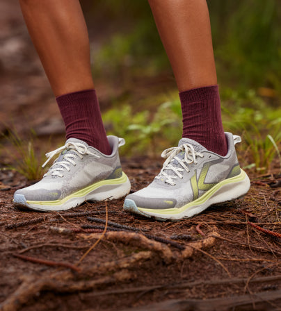 A person wearing a pair of KEEN hiking sneakers and socks on forest terrain.