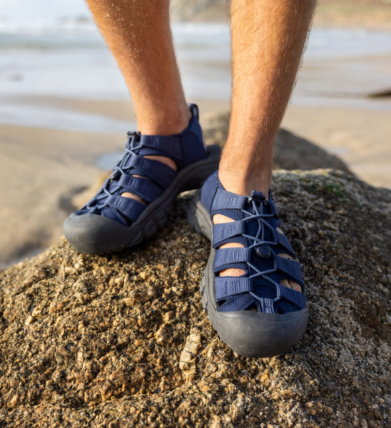 Knee-down shot of man wearing royal blue and black Newport sandals and standing on a coastal rock. 