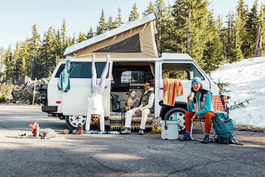A group of three people and a dog sitting around a camper van, with a winter landscape behind them.