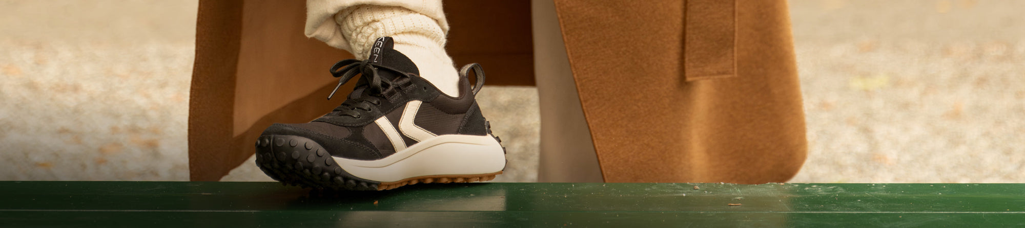 A black and white KEEN sneaker on a dark green surface, with the wearer wearing a camel-colored coat.