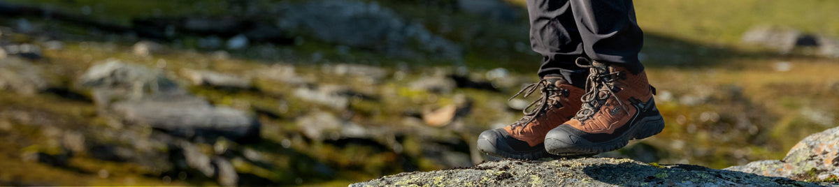 A banner image of a person standing outside on a rock, wearing a pair of brown KEEN Targhee IV backpacking boots.