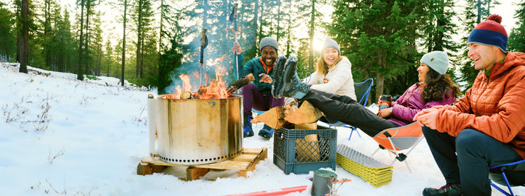 2 men and 2 women gathered around a firepit in the snow and resting their feet on a log pile