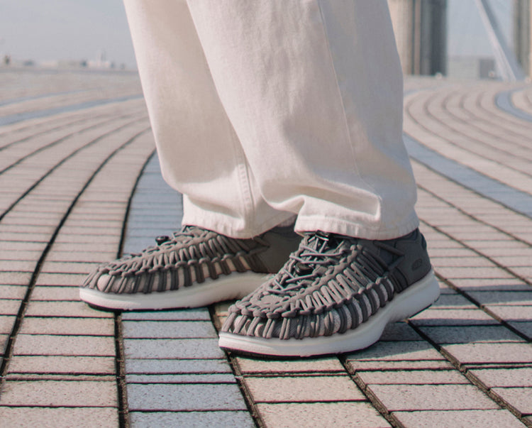 Knee down shot of man wearing gray Uneek O3 sandals and white jeans, standing on  tiled pavement.